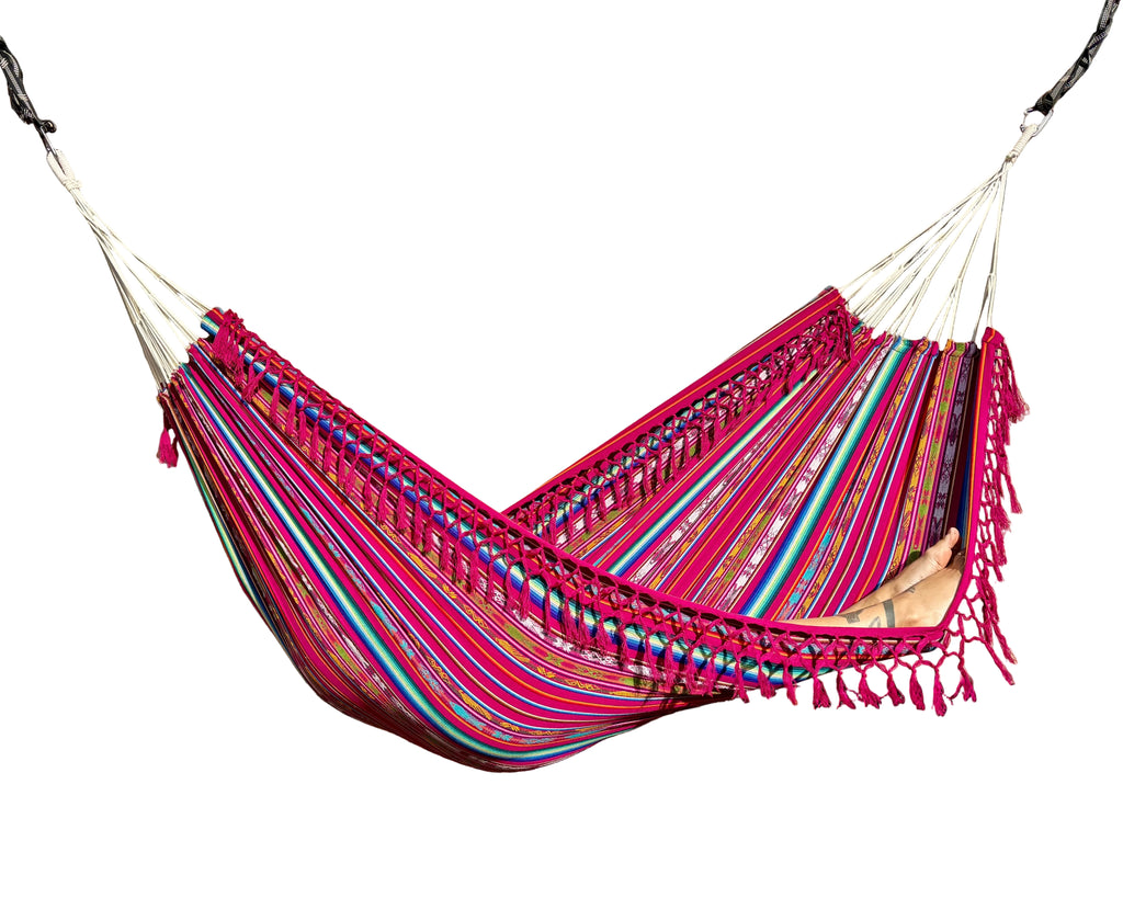 A colorful striped hammock with pink, red, blue, and green bands and fringe tassels, suspended over a wooden deck with a beige wall backdrop.