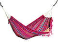 A colorful striped hammock with pink, red, blue, and green bands and fringe tassels, suspended over a wooden deck with a beige wall backdrop.